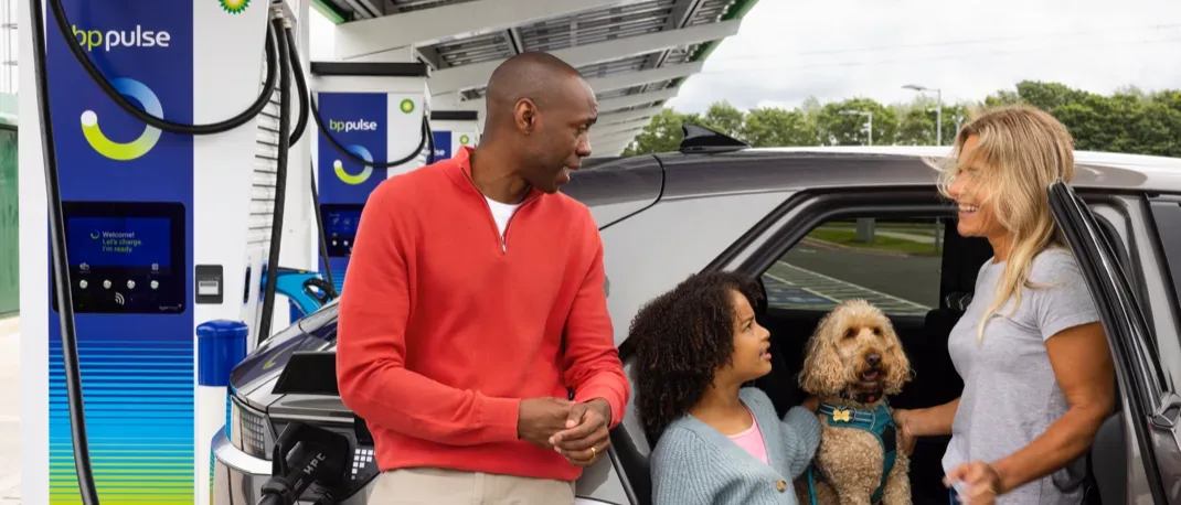 A family is standing next to their EV whilst it is plugged into a bp pulse ev charger at a bp pulse charging station. The back door of the car is ajar and the family are stood in the open door smiling at each other and petting their pet dog sat on the backseat.