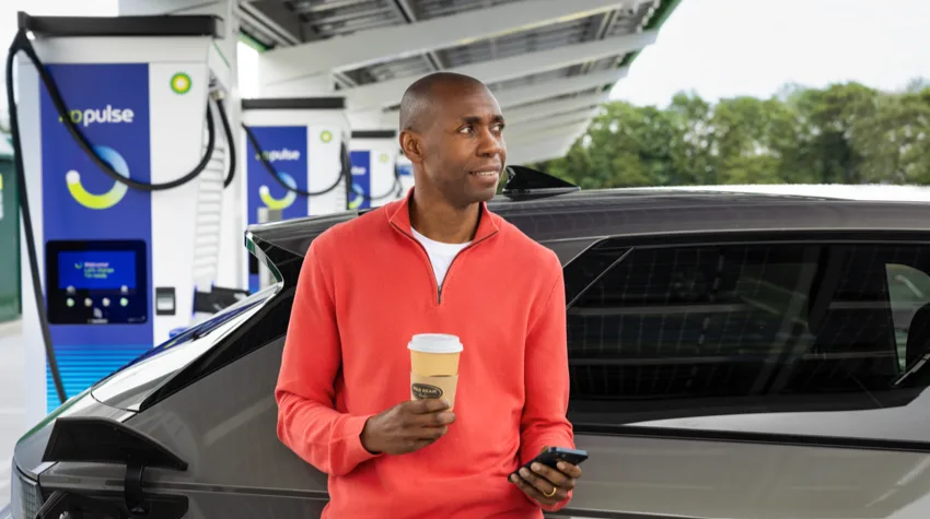 A man is drinking his coffee whilst his electric vehicle is charging at a bp pulse EV charger.