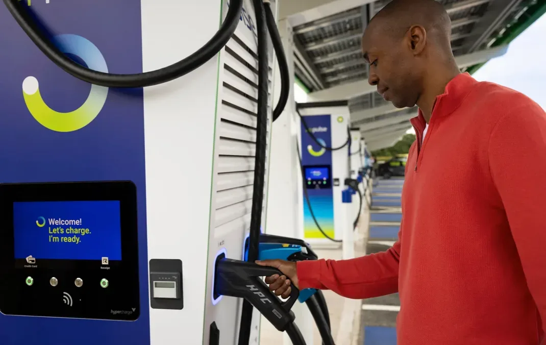 A man standing at a bp pulse charge station. He is wearing a red jumper and has his hand on the charging lead. 