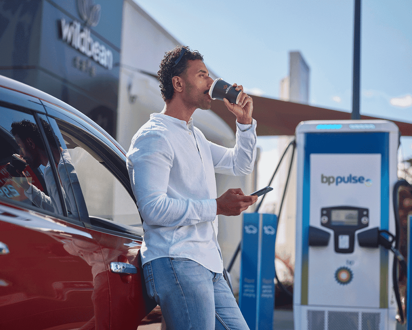 A man in a white shirt drinks his Wild Bean coffee and checks his phone whilst his electric vehicle is charging on a bp pulse charger. 