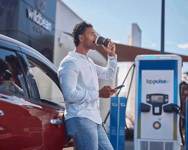 A man in a white shirt drinks his Wild Bean coffee and checks his phone whilst his electric vehicle is charging on a bp pulse charger. 