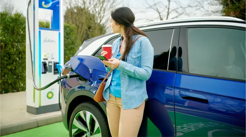 A woman holding her coffee mug, observing her electric car getting charged at a bp pulse station.