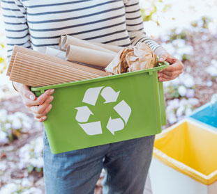A woman holding a green recycling bin filled with paper and cardboard.