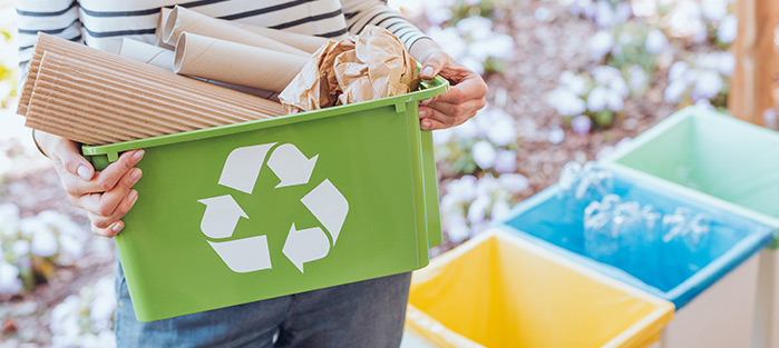 A person holding a green recycling bin filled with paper waste, standing near colorful recycling bins outdoors