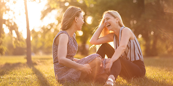 Two women laughing and enjoying a sunny day outdoors, sitting on grass surrounded by trees