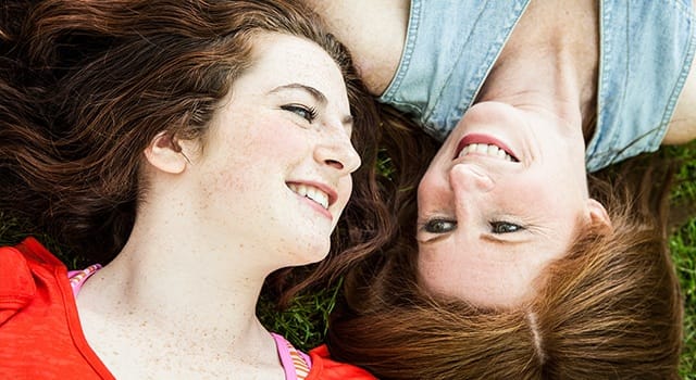 Two smiling friends lying on grass, enjoying a sunny day outdoors with hair spread around them, radiating happiness