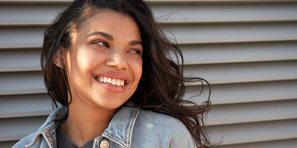 A smiling young woman with wavy hair poses against a textured gray background, radiating warmth and positivity