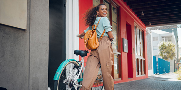 A young woman with curly hair stands next to a bicycle, smiling and wearing a casual outfit against a colorful urban backdrop