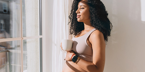 A pregnant woman with long curly black hair, standing by the window with a coffee cup