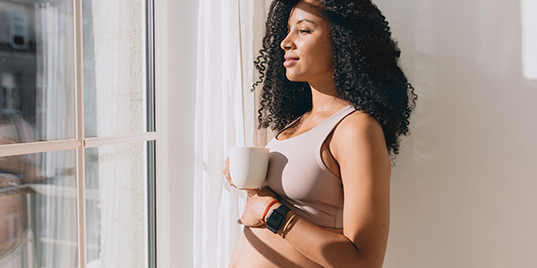 Pregnant woman with curly hair holding a coffee cup, gazing thoughtfully out a window in soft natural light