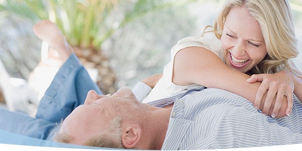 Joyful couple laughing together while lying down outdoors, surrounded by greenery, sharing a playful moment