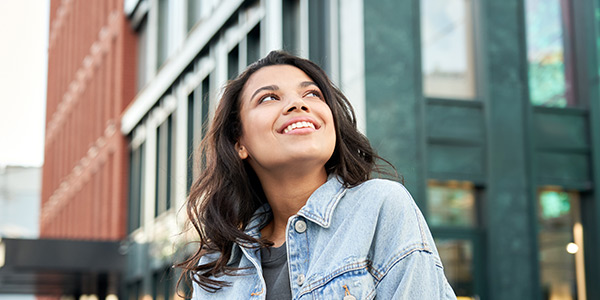 A woman with long brown hair and a jean jacket smiling in the city.