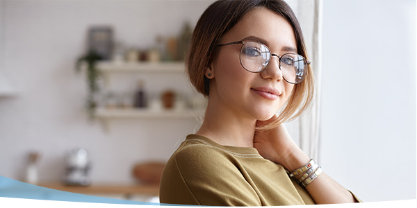 Young woman with glasses smiles warmly, showcasing a cozy indoor setting with natural light and plants