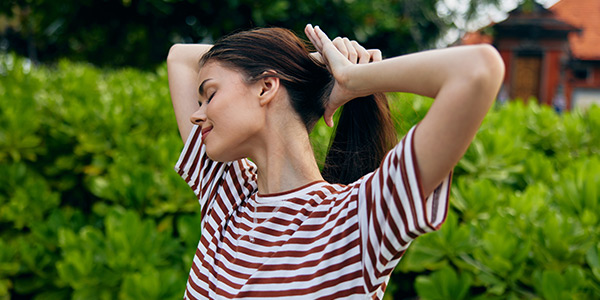 A young woman in a striped shirt ties her hair up, surrounded by lush green foliage, enjoying a sunny day outdoors