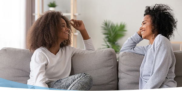 Two women with curly hair sit on a sofa, smiling and engaging in a friendly conversation in a cozy living room setting