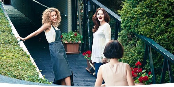 Three women enjoying a sunny outdoor gathering, smiling and interacting amidst greenery and flowers