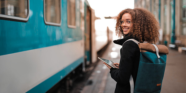 Young woman with curly hair smiles while waiting by a blue train, holding a tablet and carrying a backpack