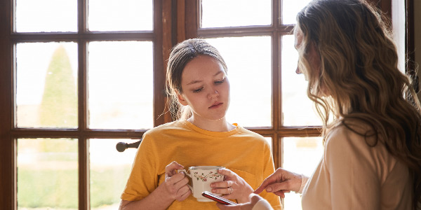 A woman with a mobile phone in her hand is talking to a girl holding a cup