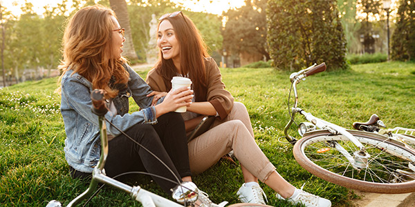 Two girls sitting on a grass field, smiling and drinking coffee