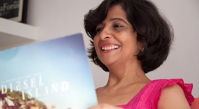 Smiling woman in a pink top reading a book titled Diesel Island, in a well-lit room with shelves