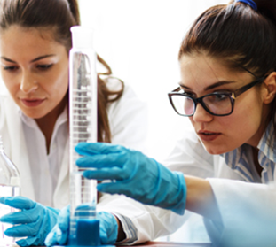 Two women in lab coats and gloves conduct an experiment with beakers and a graduated cylinder in a laboratory setting