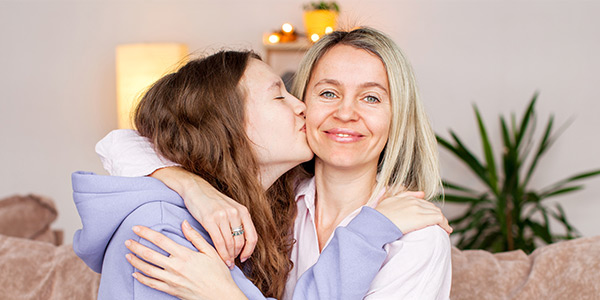 A woman smiles as a younger person kisses her cheek in a cozy living room, surrounded by greenery and warm light