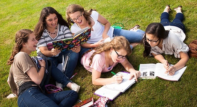 Group of five students sitting on grass, studying together with books, laughing and enjoying their time outdoors