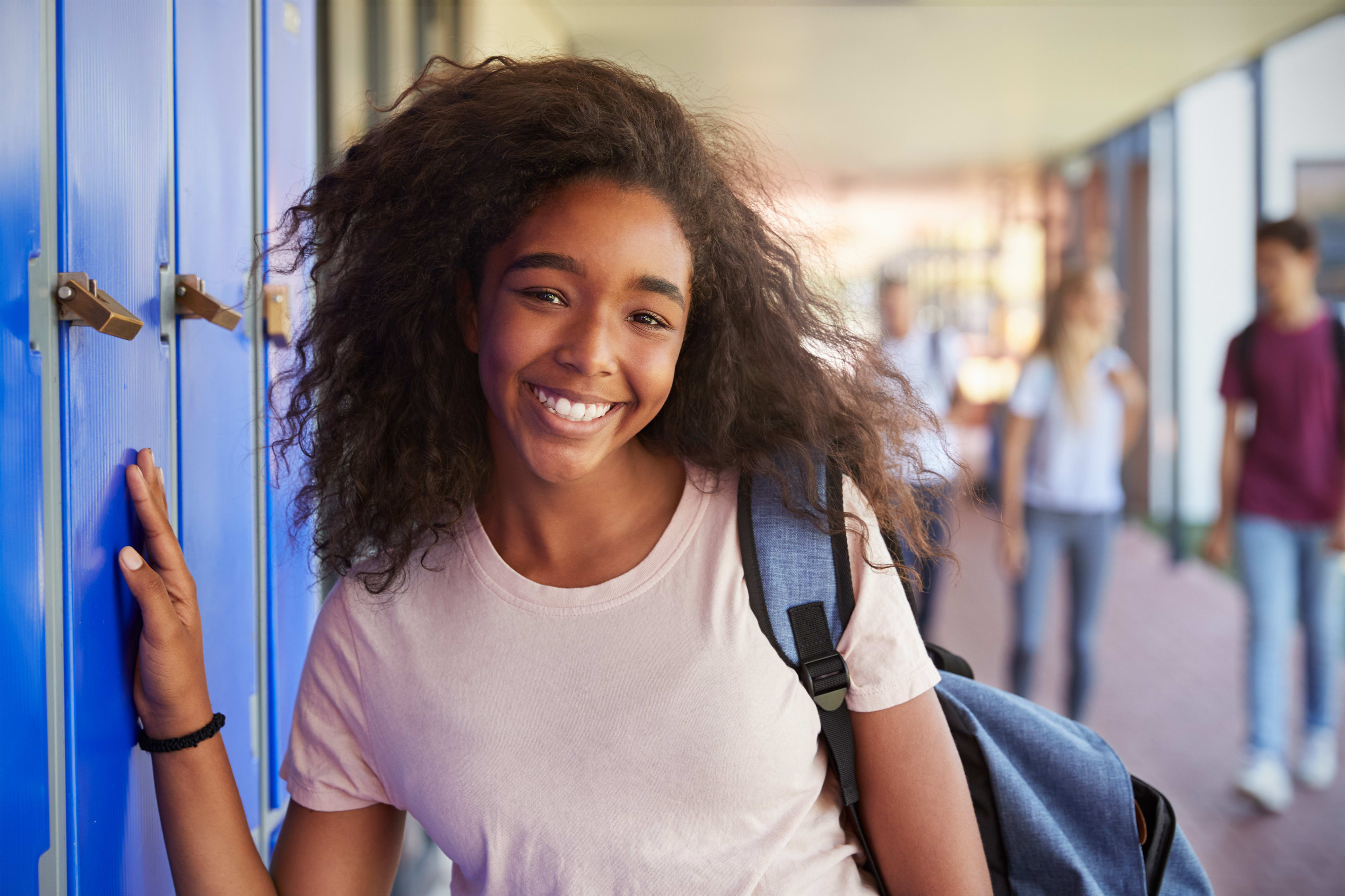 Smiling student with curly hair leaning on blue lockers, wearing a backpack, in a school hallway