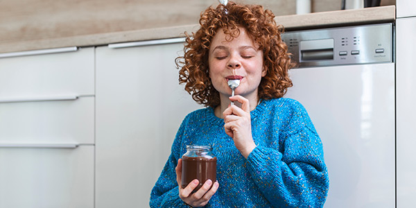 Child enjoying a spoonful of chocolate spread from a jar while sitting on the kitchen floor, smiling happily