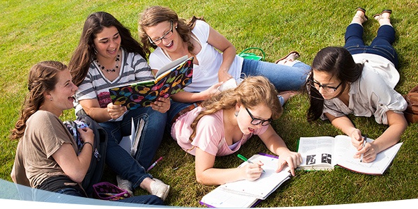 Five students sitting on grass, engaged in group study while writing and reviewing notes with books and a notebook