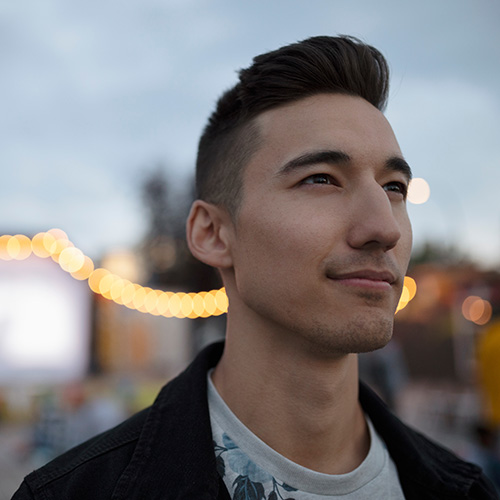 Un jeune homme aux longs cheveux fluides pose en extérieur dans une lumière naturelle chaleureuse.