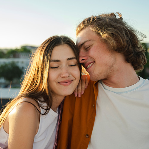 Un couple s’enlace chaleureusement sur un toit au coucher du soleil, tous deux avec des cheveux bien coiffés.