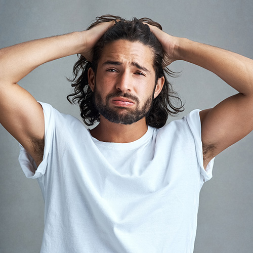 Un homme barbu en t-shirt blanc joint les mains derrière la tête en regardant vers le haut.