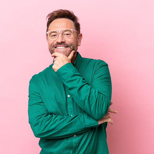 Un homme barbu en chemise verte sourit largement sur un fond de studio rose.