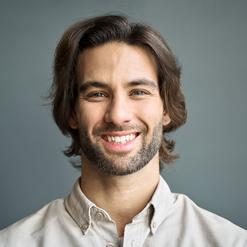 Un homme souriant avec de longs cheveux foncés d’apparence saine, portant une chemise blanche.