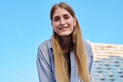 Une jeune femme aux longs cheveux blonds sourit face à l’objectif, sur un fond bleu uni.