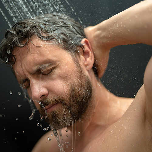 Un homme barbu fait mousser du shampoing dans ses cheveux sous la douche, les yeux fermés.
