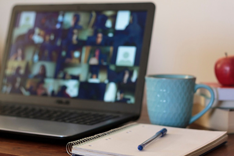 A pen, paper and mug of tea or coffee on a desk with a laptop showing a busy online meeting in the background