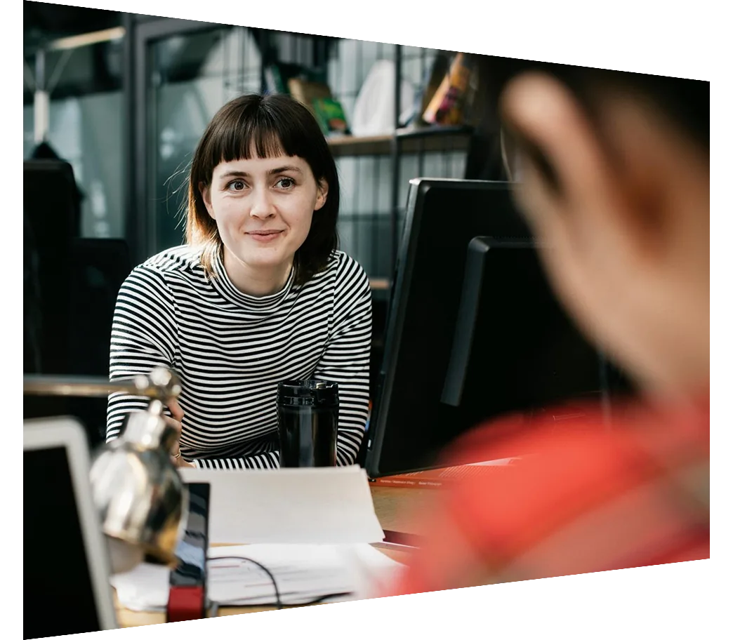 A young woman sitting at her desk and talking to one of her colleagues while working in a modern office space.