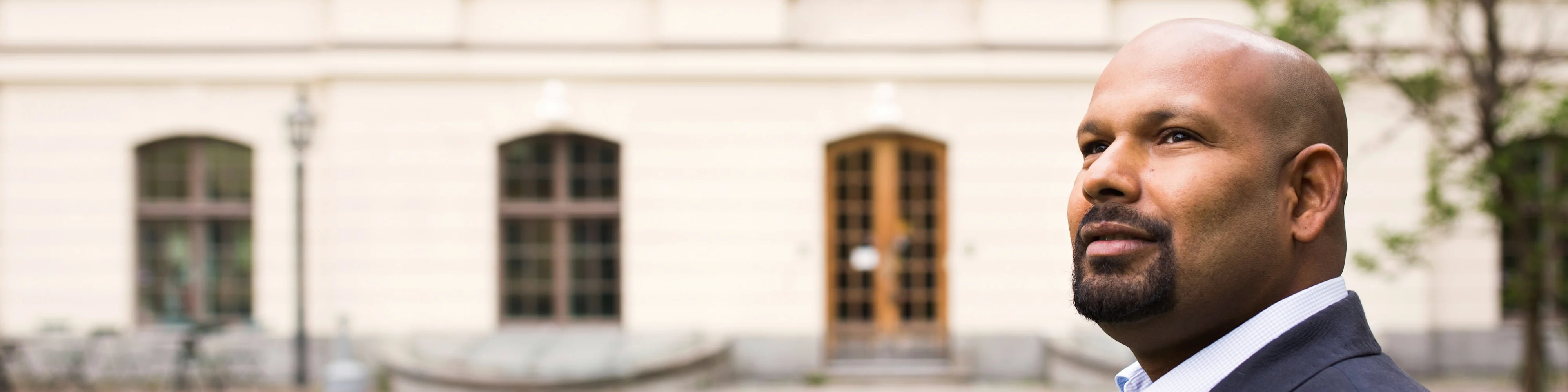 A black male in a suit in front of a university campus building looking off into the distance with a proud and hopeful expression. 