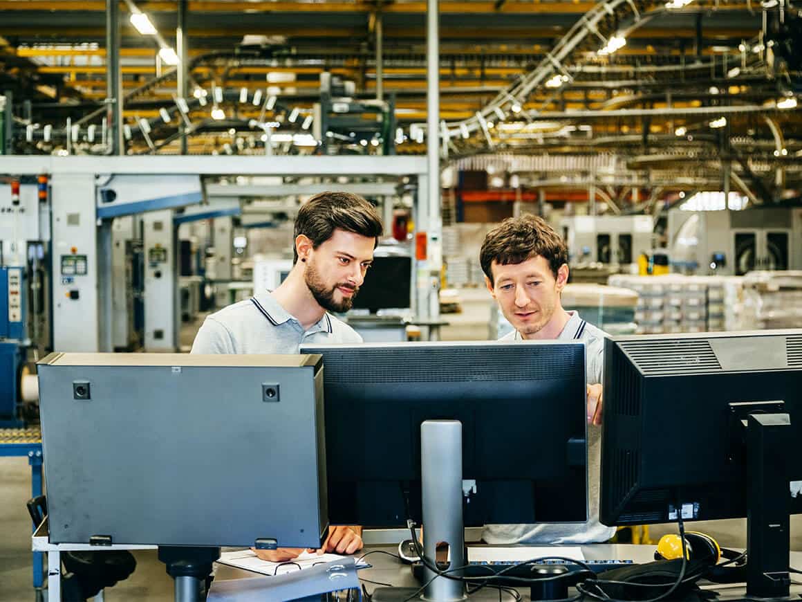 Two engineers standing behind computer monitors in a large factory