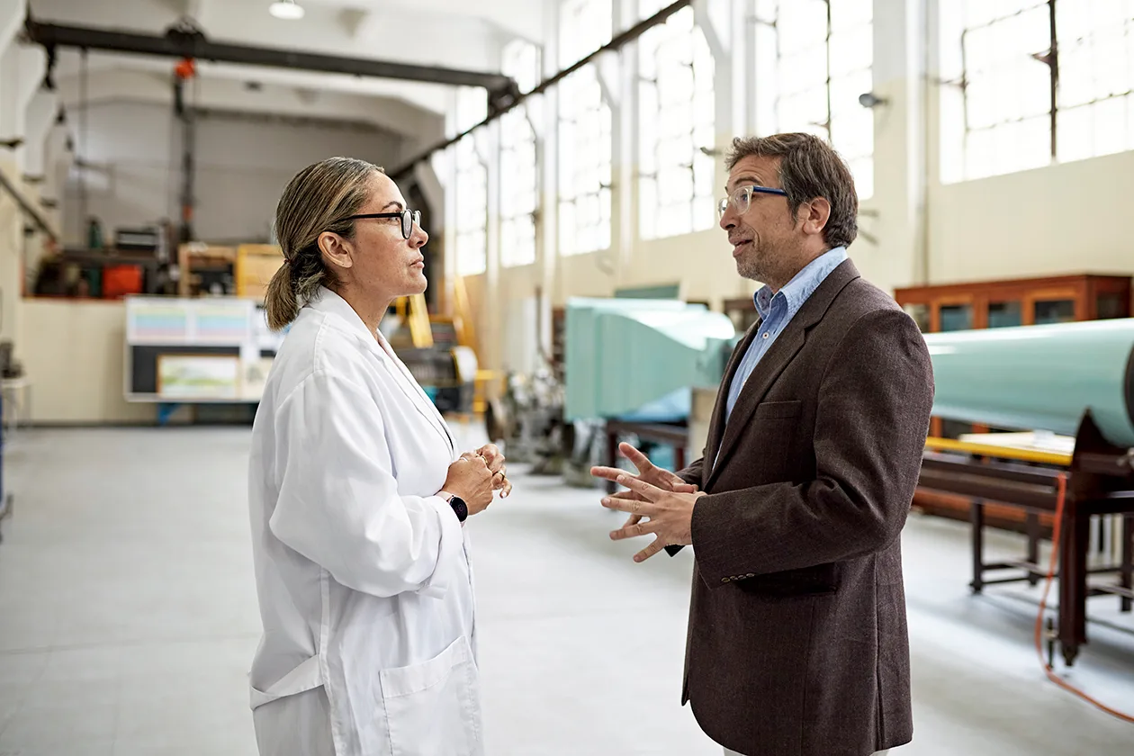 Two people at an industrial facility speaking. One is wearing a lab coat and googles, the other is wearing casual business attire. 