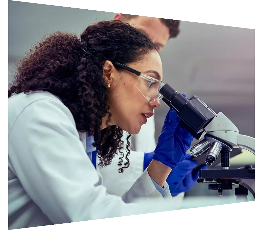 Female scientist wearing safety goggles and gloves, focusing on analyzing test sample under microscope, observed by male colleague in laboratory