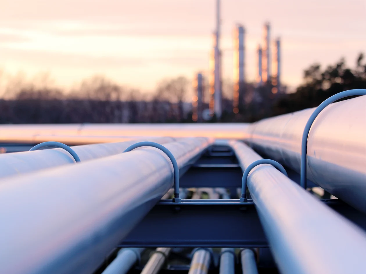 A series of pipes in the foreground with an oil refinery and a sunset in the background