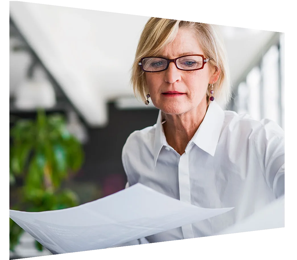 A woman in glasses reviews documents with focus, surrounded by paperwork.