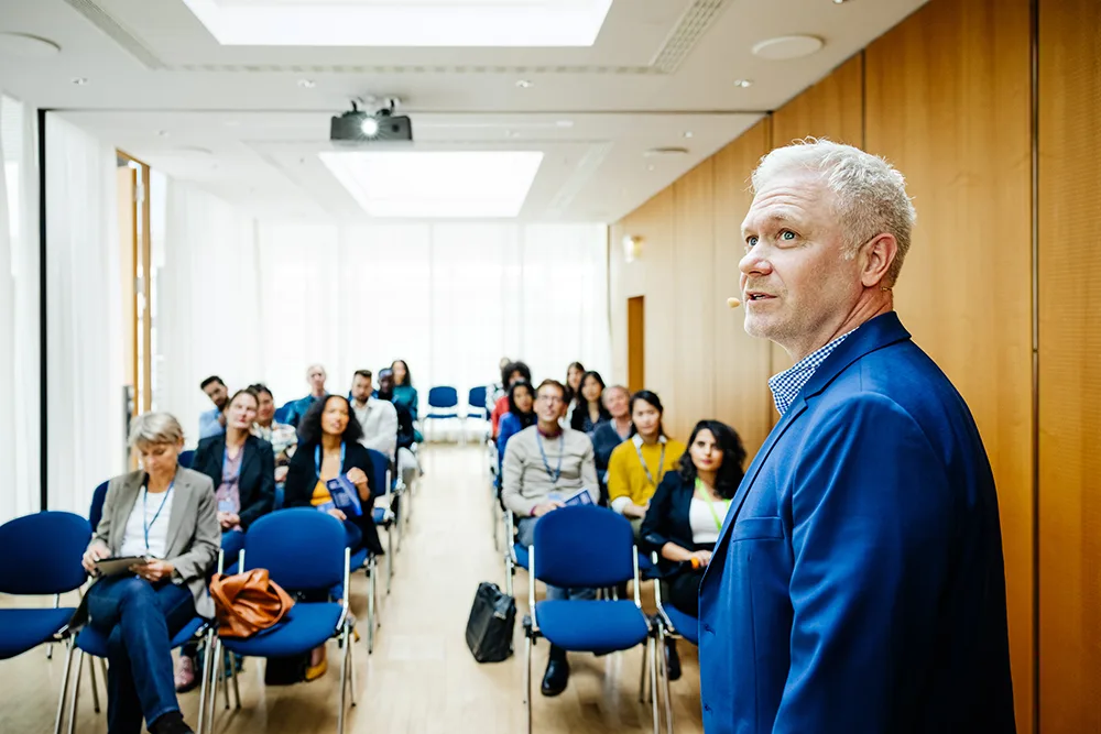 A man with gray hair and in a blue sports coat is looking off towards a screen (unseen) and presenting to an audience of peers.
