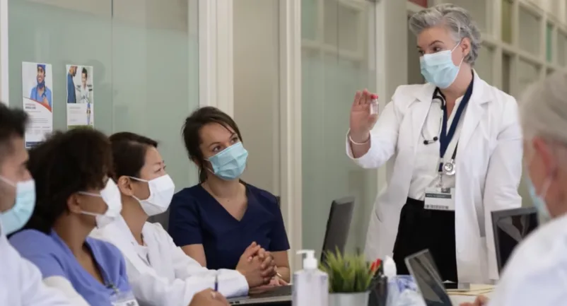 Female physician wearing mask speaking in front of board room 