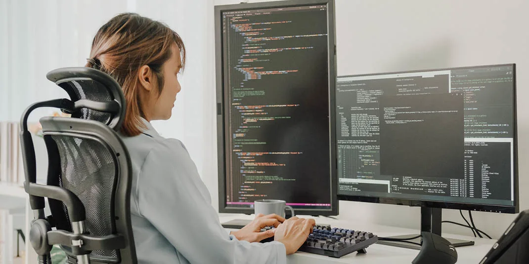 Photo of a woman at a computer with multiple screens displaying code (© istock.com/MTStock Studio)