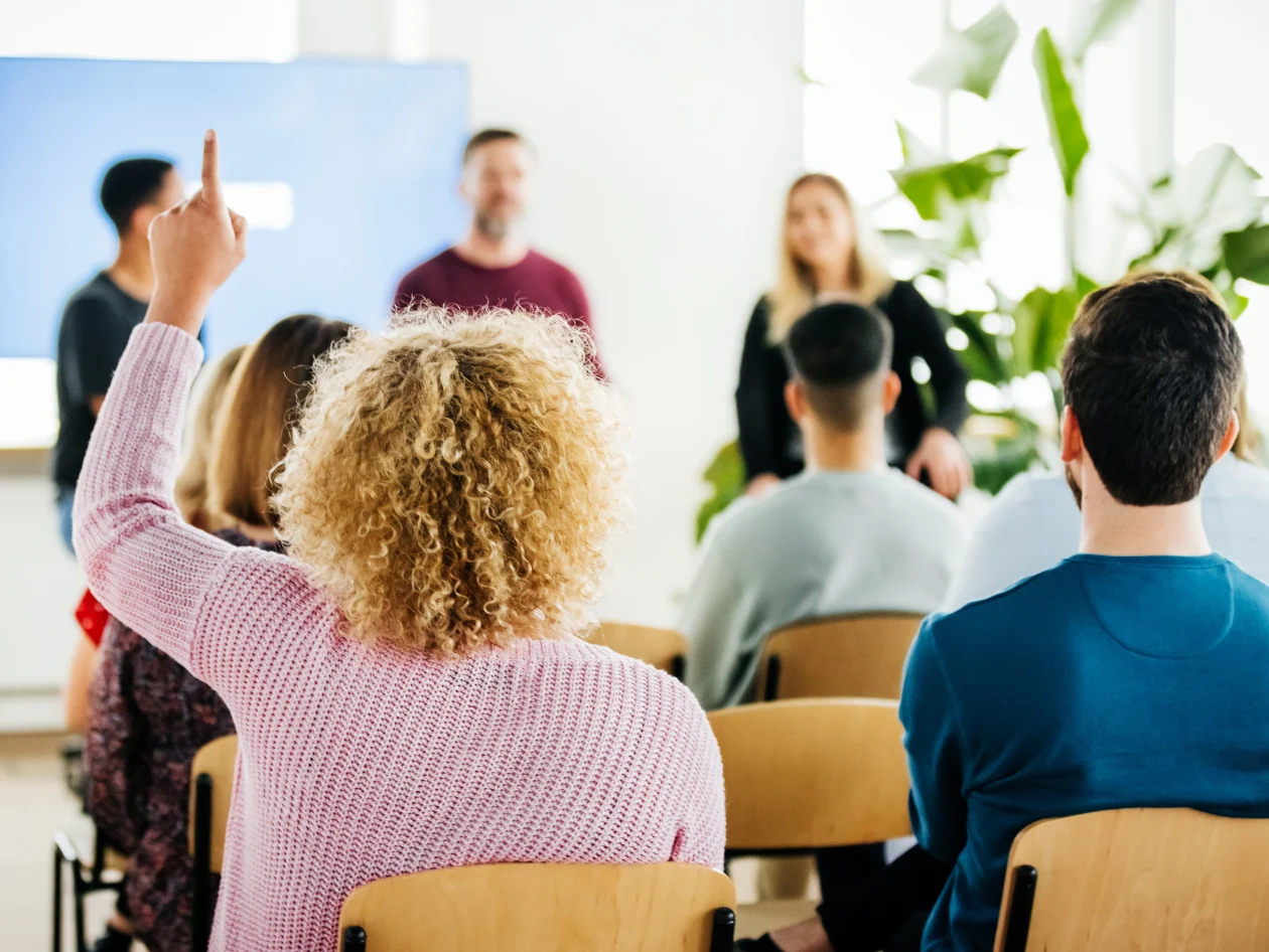 A presentation occurring with a woman raising her hand in the audience.