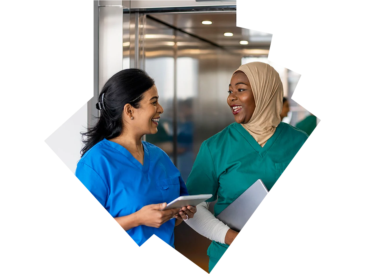 Two female nurses smiling and talking while walking by an elevator